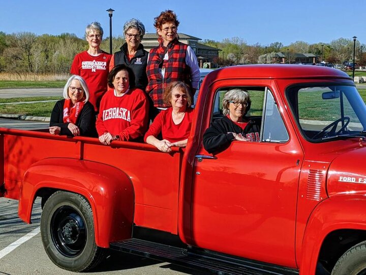 Women pose in the bed of a classic truck painted Husker red.