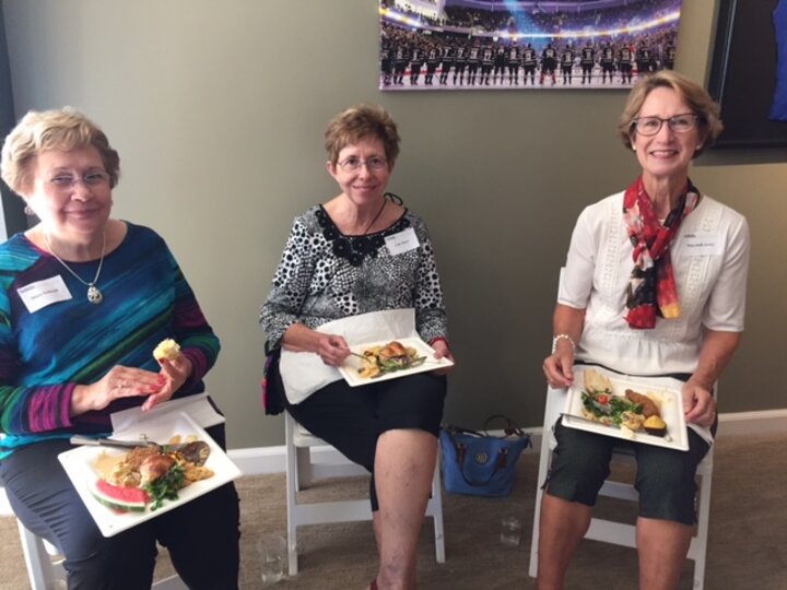 Three women sitting on folding chairs with plates of food smile at camera.