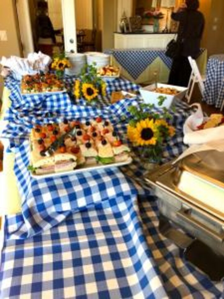 Blue and white checkered table cloth covered table filled with food garnished with sunflowers.