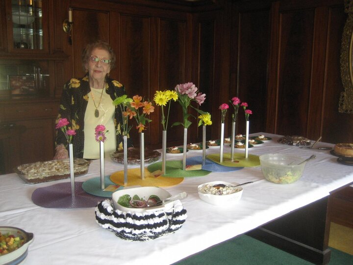 A woman poses at table with food set out with many flower bouqeuts along center of table.