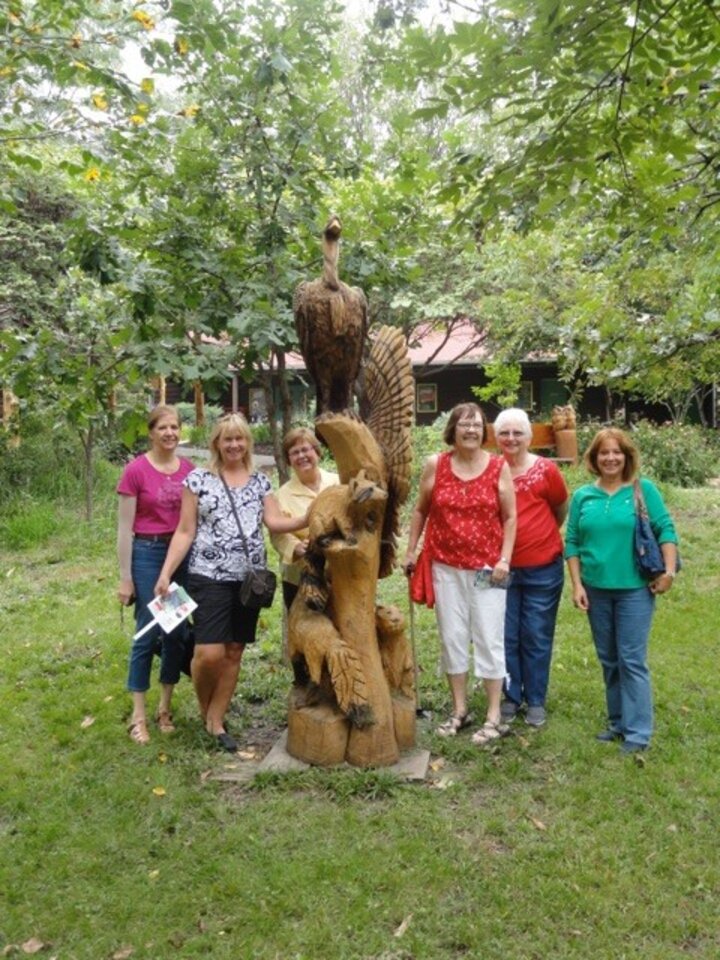 Group of women pose around large wood carving of different animals.