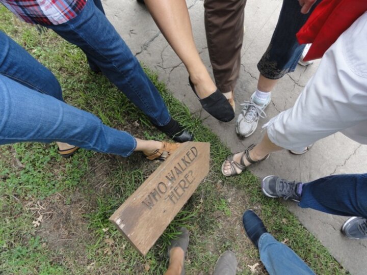 A sign reading Who Walked Here points at a spot on the ground where a large group points a foot at.