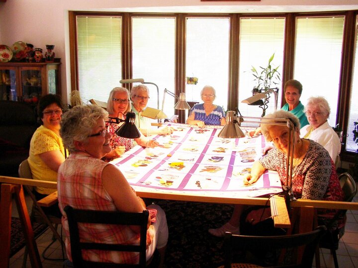 Club members sit around a large table working on a quilt.